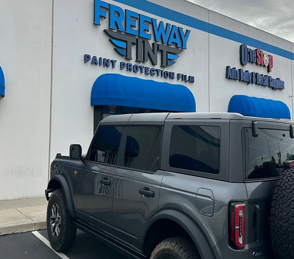 A dark gray SUV with tinted windows parked in front of the Freeway Tint storefront, which features blue awnings and signage reading "Freeway Tint Paint Protection Film."
