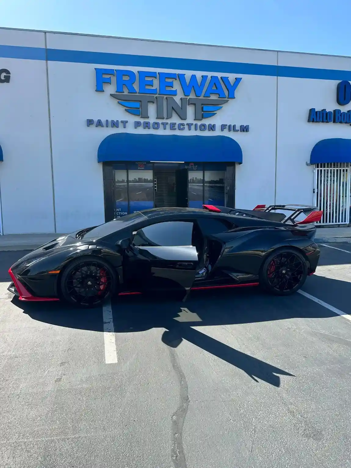 Black Lamborghini with red accents and tinted windows parked in front of Freeway Tint, with the driver's door open under the storefront sign reading “Paint Protection Film.”