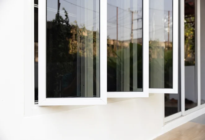 Three-panel white-framed window slightly opened outward, with tinted glass reflecting outdoor greenery and power lines, providing privacy while allowing light.