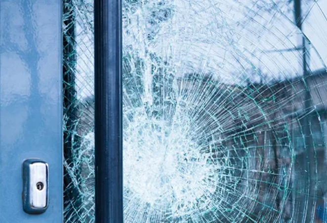 Close-up of a shattered glass door with a central impact point, showing spiderweb-like cracks, suggesting the importance of security film or reinforced glass.