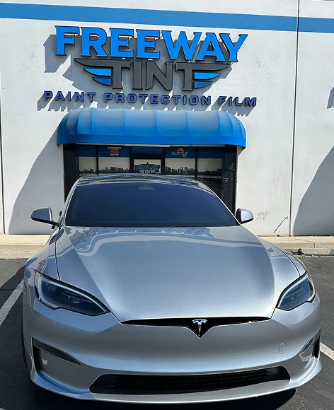 Front view of a silver Tesla Model S with dark tinted windows parked in front of the Freeway Tint storefront, which displays a blue awning and "Paint Protection Film" signage.