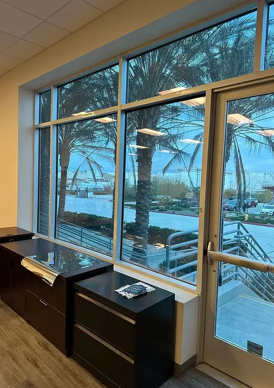 Interior view of an office space with large tinted windows and a glass door, showing palm trees and a parking lot outside.