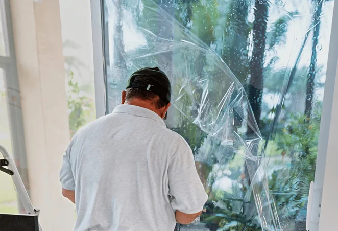 Man wearing a gray polo shirt and cap installing clear window film on a large glass door, with water and bubbles visible beneath the film during application.
