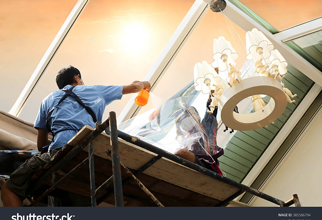 Worker standing on scaffolding applying window film to a large slanted skylight, using a spray bottle for installation near an ornate ceiling light fixture.