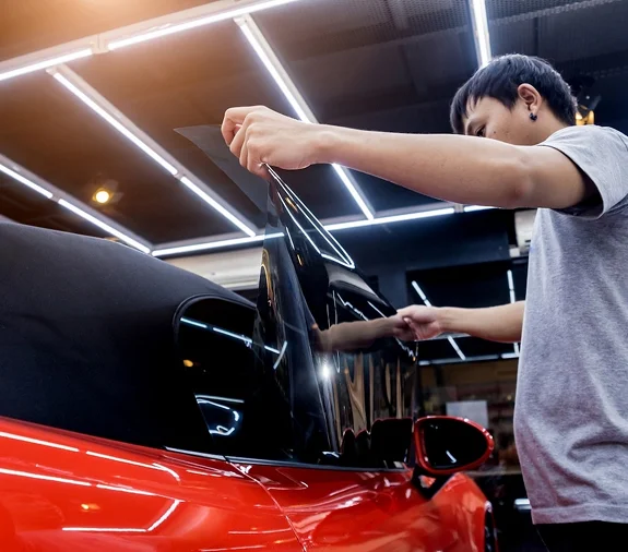 Technician applying dark window tint film to the side window of a red convertible inside a well-lit garage.