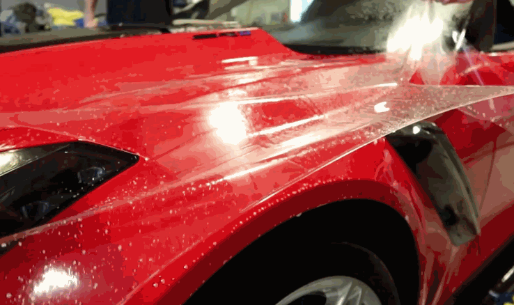 A red Corvette hood gets sprayed down as a clear paint protection film is carefully applied and smoothed over the surface.