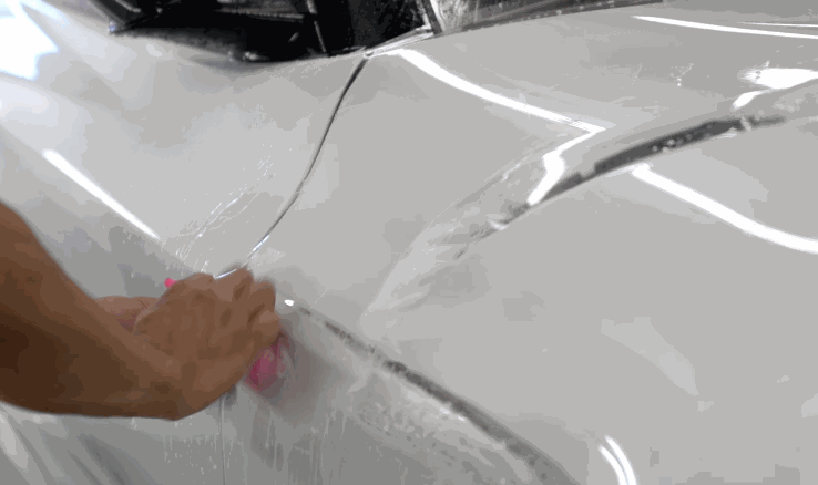 A technician uses a squeegee to press paint protection film onto the rear panel of a white sports car, smoothing out bubbles.