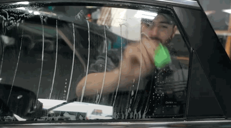 A man applies window tinting film to a car window, using a green squeegee to smooth out water and air bubbles.