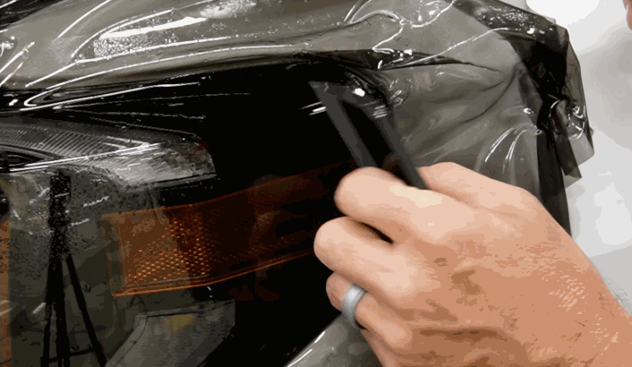 Technician using a squeegee to install paint protection film over a vehicle headlight and amber turn signal. The wet paint protection film is shaped tightly around the edges to create a seamless protective barrier.