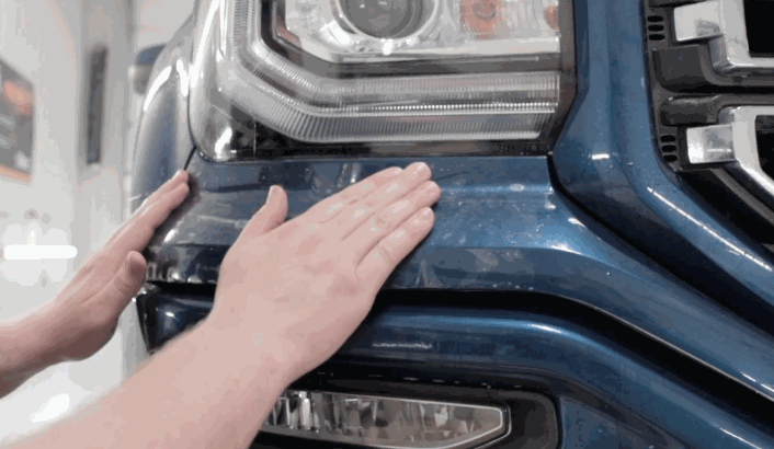 Hands pressing paint protection film onto the front bumper of a blue truck beneath the headlight. The clear paint protection film is smoothed along the curved surface to guard against rock chips and road debris.