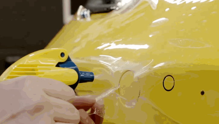 A heat gun is used to soften paint protection film on a yellow car panel as it is shaped around a circular detail. The warmed film becomes more flexible allowing it to conform tightly without wrinkles.
