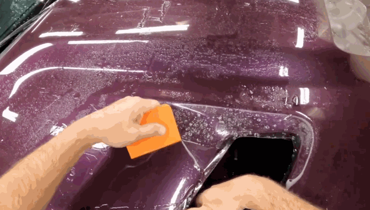 Hands press an orange squeegee across a wet sheet of paint protection film on a maroon car panel. The film is carefully stretched and flattened around a curved opening to ensure full coverage and durability.
