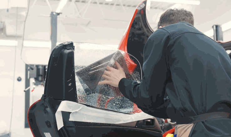 A technician carefully applies window tint film to a car door window, using hands to smooth the wet film across the glass inside a workshop. The process shows how window tint is installed to improve heat rejection and comfort.