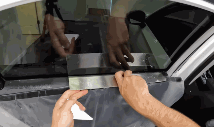 Two hands align and position window tint film along the bottom edge of a car window while moisture is visible beneath the surface. The film is carefully placed to prepare for smoothing and sealing.