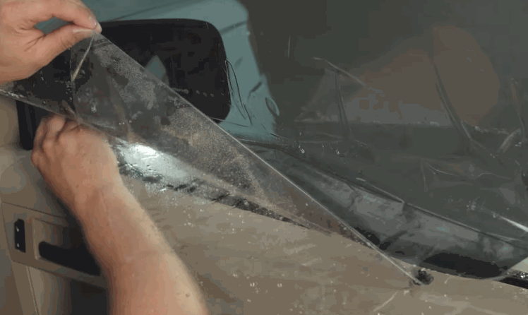 Installer peels back window tint film from a wet car window, revealing water droplets and adhesive as the film is positioned for installation. The process shows careful alignment before final application.