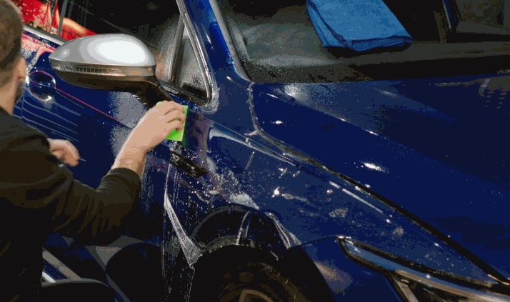 Technician applies paint protection film to the front fender and door area of a blue car using a green squeegee. Soapy water and a blue towel on the windshield show the film is being positioned and smoothed during installation.