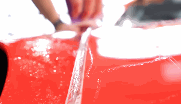 A technician stretches paint protection film across a wet red vehicle panel. The clear film is being aligned along the body line for precise coverage.