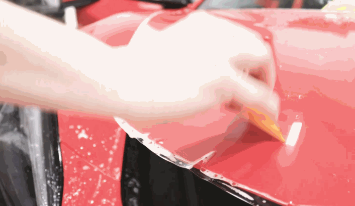A technician pulls clear paint protection film across the curved panel of a red vehicle. Water droplets on the surface help position the film for a smooth fit.