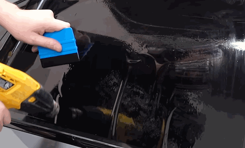 A technician uses a blue squeegee and yellow heat gun to shape ceramic window tint on a wet car window. The film is being smoothed across the glass for a clean fit.