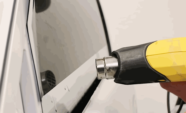 A yellow heat gun warms ceramic window tint along the edge of a light colored vehicle window. The close up shows the film being shaped to fit the glass.