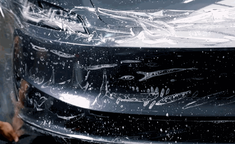 Close-up of paint protection film stretched across the front bumper and headlight area of a black car.