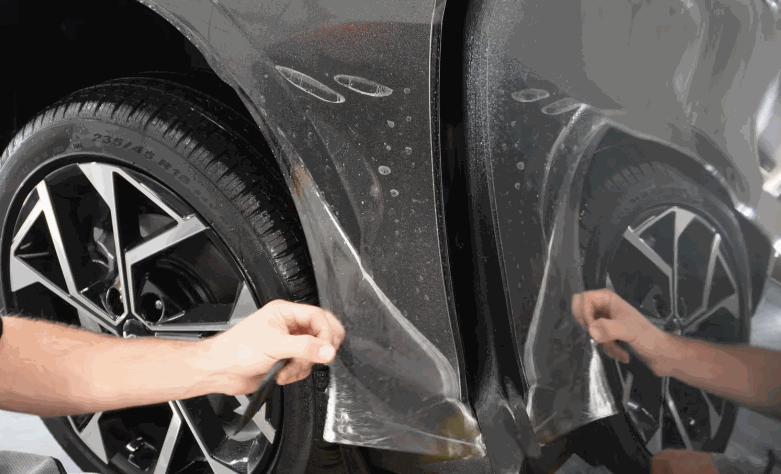 Technician pulls paint protection film around the wheel arch and lower side panel of a gray car.