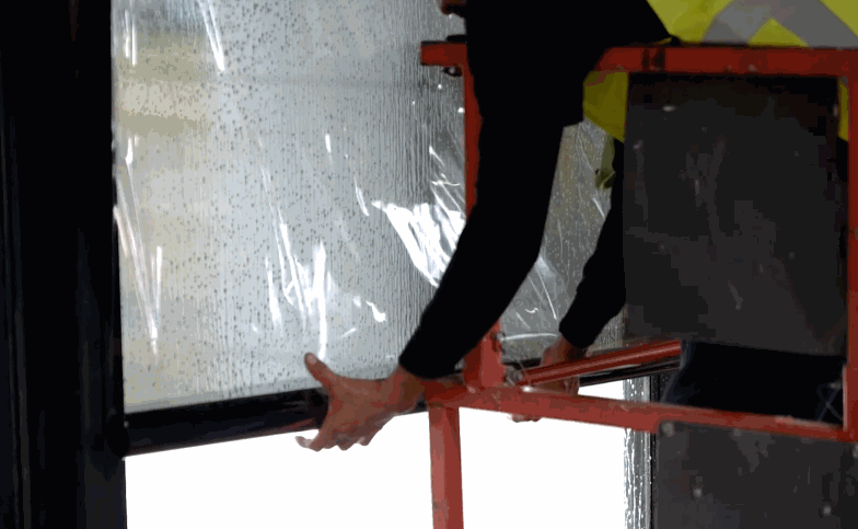 Installer on a lift smooths commercial window tint across a large glass panel with water droplets on the surface.