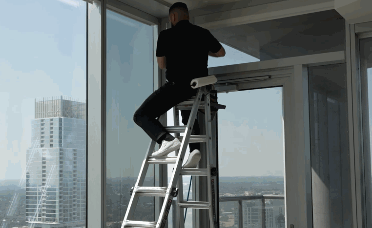 Installer works from a ladder applying commercial window tint to tall glass windows in a high rise building.
