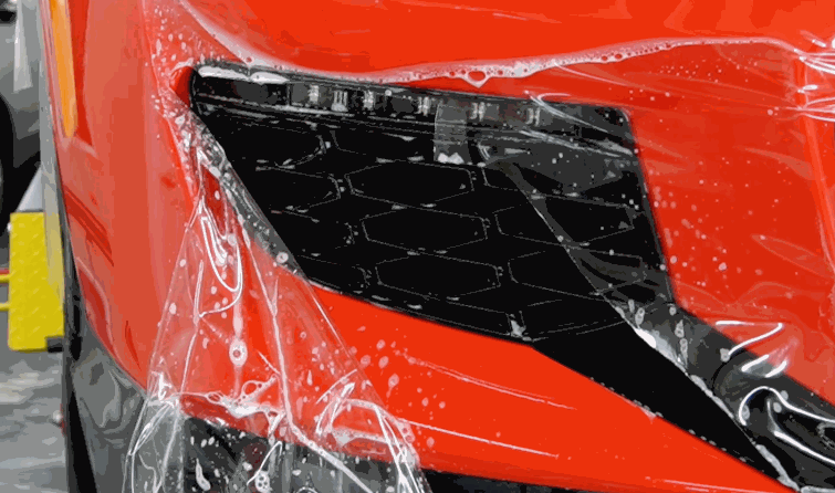 Paint protection film applied around a black grille opening on the front bumper of a red car with installation solution visible across the surface.