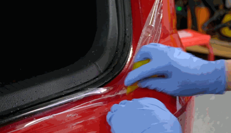 Technician wearing blue gloves trims clear paint protection film along the edge of a red vehicle near a black window seal.