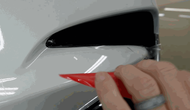 A technician uses a red detailing tool around a recessed area on a white vehicle panel. Careful edge work helps create a clean finish before ceramic coating is applied over paint protection film.